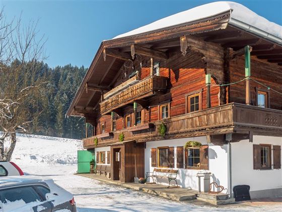 A traditional wooden house in the snow, surrounded by snow-covered trees. The sky is clear and the sun is shining.