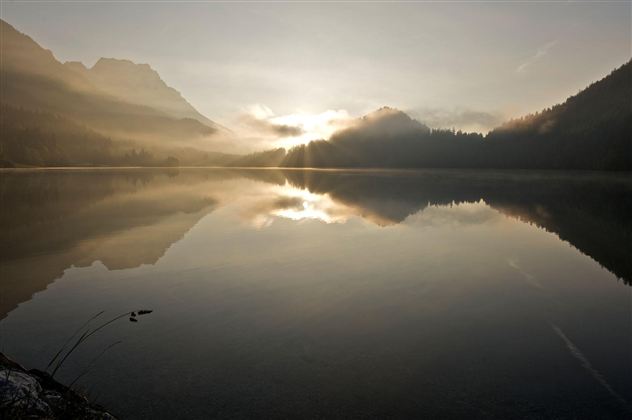 Ein ruhiger See mit spiegelndem Wasser und sanften Bergformen im Hintergrund. Die Morgensonne strahlt durch den Nebel und schafft eine friedliche Atmosphäre.