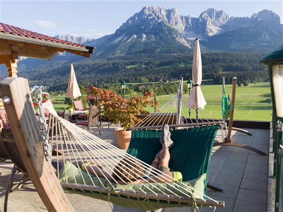 A terrace with a hammock and a view of the mountains. The surroundings are green and inviting, perfect for relaxing.