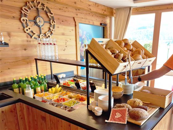 Een buffet met verse broodjes, fruit en verschillende dranken. De houten bekleding en het grote raamgebied creëren een uitnodigende sfeer.