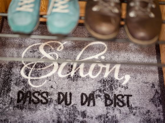 Een welkommat met de tekst "Fijn dat je er bent". Bovenop zijn een paar schoenen in verschillende kleuren te zien.