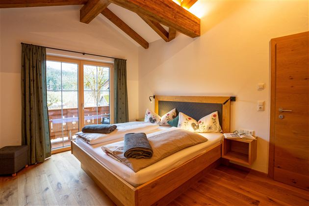 A cozy bedroom with a double bed and wooden furniture. Large windows offer views of the balcony and nature.