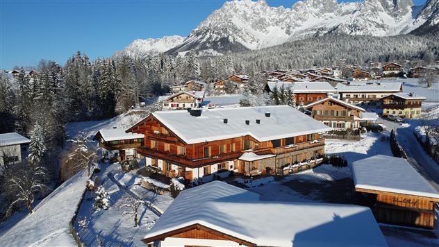 Eine malerische Alpenlandschaft mit schneebedeckten Bergen. Im Vordergrund befinden sich charmante Holzhäuser in einer ruhigen, winterlichen Umgebung.