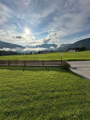 Eine grüne Wiese mit einem schönen Blick auf die Berge und einen bewölkten Himmel. Die Sonne scheint durch die Wolken und schafft eine friedliche Atmosphäre.
