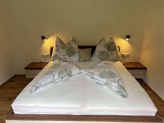 Ein gemütliches Schlafzimmer mit einem großen Bett und dekorativen Kissen. Die Wände sind neutral, und es gibt sanfte Beleuchtung an beiden Seiten des Bettes.
