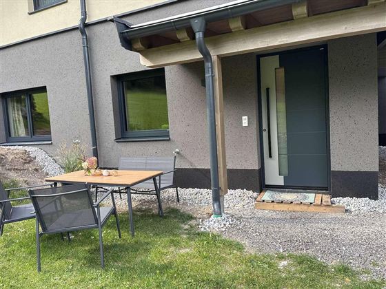 Eine moderne Haustür mit einem kleinen Vorplatz und einem Tisch mit Stühlen daneben. Der Bereich ist von einem gepflegten Garten umgeben.
