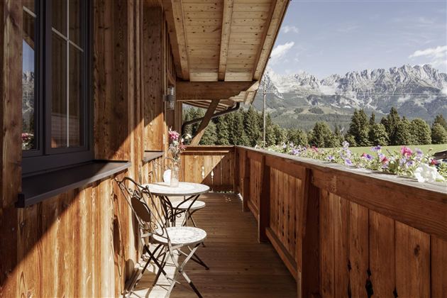 A cozy wooden balcony with a small table and two chairs. In the background, impressive mountains and green forests can be seen.