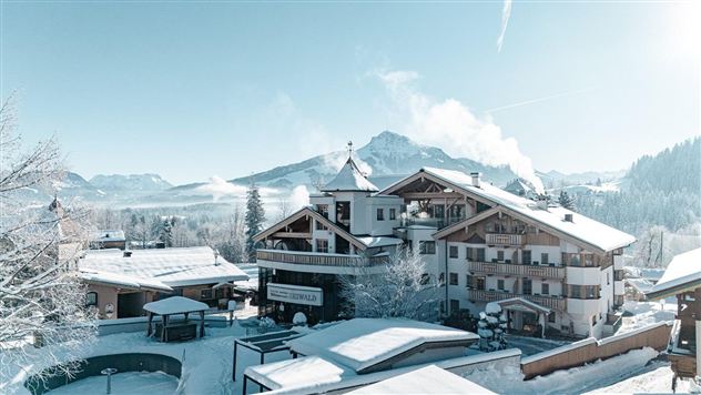 Eine verschneite Landschaft mit einem gemütlichen Hotel und schneebedeckten Bergen im Hintergrund. Der klare Himmel zeigt eine ruhige Winteratmosphäre.