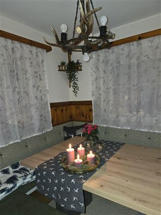 Ein gemütlicher Essbereich mit einem Holztisch, dekoriert mit Kerzen und einer kleinen Pflanze. Die Fenster sind mit weißen Vorhängen geschmückt, und ein rustikaler Kronleuchter hängt von der Decke.