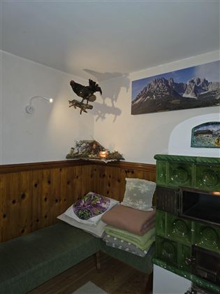 Ein gemütlicher Raum mit einer Holzbank und bunten Kissen. An der Wand hängt ein Bild von Bergen, während ein grüner Ofen in der Ecke steht.