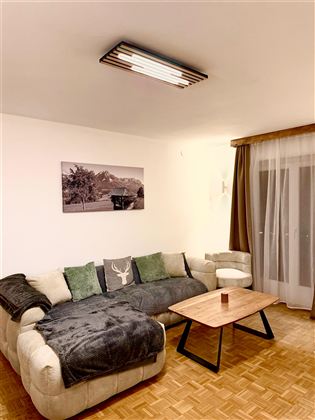Ein gemütliches Wohnzimmer mit einem großen Sofa, einem Couchtisch und einem Bild an der Wand. Die Fenster sind mit Vorhängen dekoriert, und der Raum hat warmes Licht.