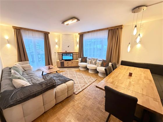 Ein modernes Wohnzimmer mit einer bequemen Couch, einem Tisch und einem Fernseher. Große Fenster sorgen für viel natürliches Licht.