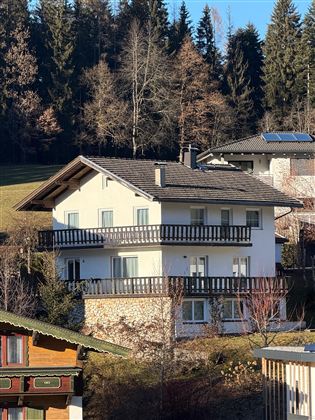 Ein modernes Haus mit mehreren Balkonen inmitten einer grünen Landschaft. Im Hintergrund sind hohe Bäume und weitere Wohngebäude sichtbar.