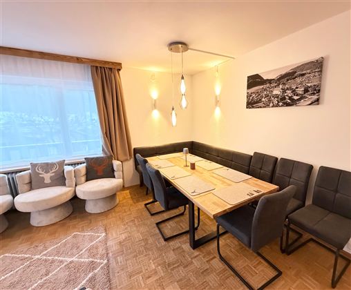 Ein modernes Esszimmer mit einem großen Holztisch und bequemen Stühlen. An der Wand hängt ein schwarzes und weißes Bild, und das Fenster bietet viel Tageslicht.