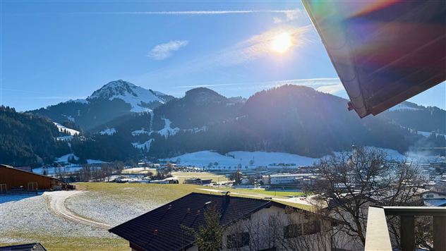 Eine malerische winterliche Landschaft mit schneebedeckten Bergen und einem klaren blauen Himmel. Die Sonne scheint über die sanften Hügel und die grünen Wiesen.