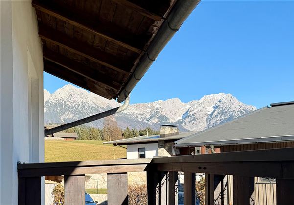 Eine schöne Aussicht auf schneebedeckte Berge unter einem klaren blauen Himmel. Im Vordergrund ist ein Balkon mit Holzgeländer sichtbar.