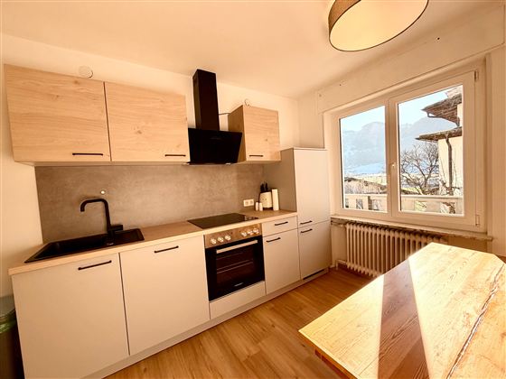 Eine moderne Küche mit Holzschränken und einem kompakten Design. Die große Fensterfront sorgt für viel Tageslicht und einen Blick ins Freie.