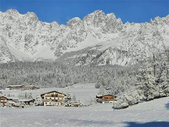 Eine verschneite Landschaft mit Bergen und einem blauen Himmel. Im Vordergrund sind einige charmante Hütten zu sehen.