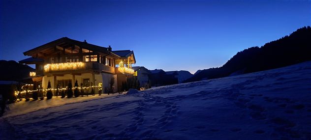 Ein gemütliches Holzhaus im Schnee, beleuchtet von warmem Licht. Der Himmel ist klar und zeigt einen schönen, tiefblauen Farbton.