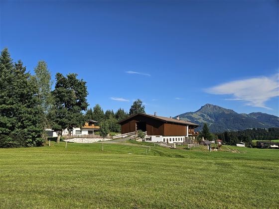 Eine malerische Landschaft mit einem großen Haus und grünen Wiesen. Im Hintergrund erstrecken sich Berge unter einem klaren blauen Himmel.