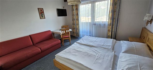 Ein gemütliches Hotelzimmer mit einem Doppelbett, einem Sofa und einem Tisch. Große Fenster bieten viel Tageslicht und eine angenehme Atmosphäre.