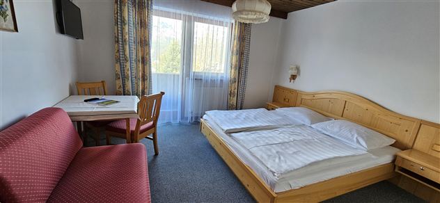 Ein einfaches Hotelzimmer mit zwei Betten und einem Tisch. Es gibt einen Wohnbereich mit einem Sofa und einen Balkon, der viel Tageslicht hereinlässt.