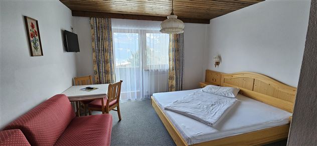 Ein gemütliches Zimmer mit einem großen Bett, einem Tisch und Stühlen. Große Fenster sorgen für viel natürliches Licht.