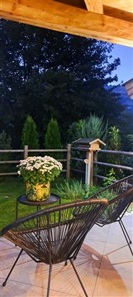 A cozy terrace with two chairs and a small table. On the table is a pot of flowers, surrounded by green garden.