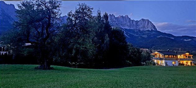 A calm landscape with a large tree and a house in the foreground. In the background, impressive mountains rise under an evening sky.