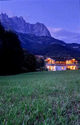 A stylish house in the mountains at twilight, surrounded by meadows. The warm lights of the house shine into the peaceful landscape.