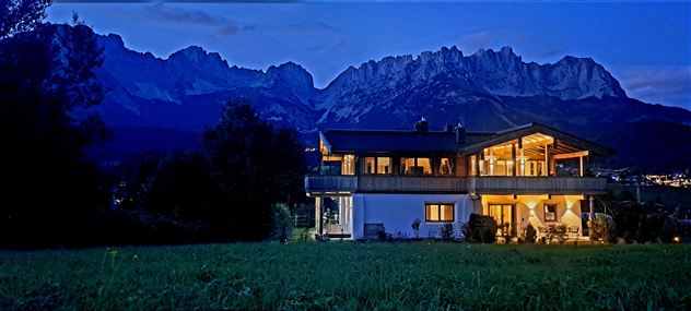 A modern house at dusk, surrounded by majestic mountains. The soft lights of the house shimmer in the serene landscape.