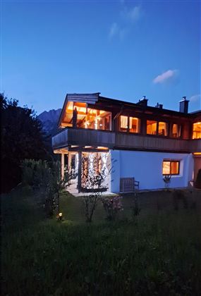 A modern house with cozy lights that shines in the twilight. In the background, gentle hills and a clear evening sky can be seen.