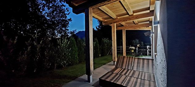 A cozy porch with a wooden ceiling under a clear night sky. Nearby, plants and trees are visible.