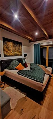 A cozy bedroom with a large bed and modern pillows. The wooden beam ceiling and the artistic picture on the wall give the room a warm atmosphere.