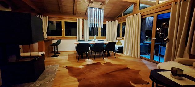 A modern living room with wooden floors and large windows. The space is elegantly furnished and features a dining area with a table and chairs.