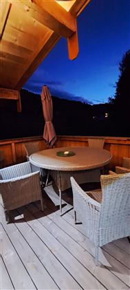 A cozy outdoor area with a round table and chairs made of rattan. The sky is dark blue, indicating the evening.