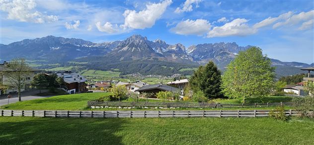A picturesque landscape with green meadows and an impressive mountain range in the background. The sky is clear and blue with some clouds.