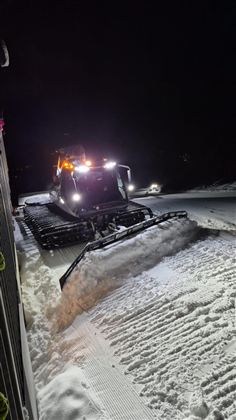 Ein Pistenraupe arbeitet nachts auf einem schneebedeckten Weg. Die Lichter des Fahrzeugs leuchten in der Dunkelheit.