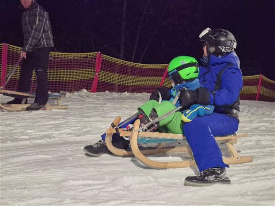 Ein Erwachsener zieht einen Schlitten auf einer Schneebahn. Auf dem Schlitten sitzt ein Kind, beide tragen Winterkleidung und Schutzhelme.