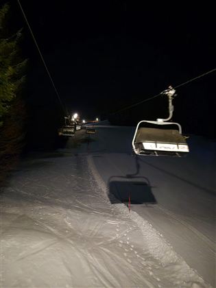Eine Skiliftanlage bei Nacht auf einer verschneiten Piste. Die Umgebung ist düster, mit wenigen Lichtquellen in der Ferne.