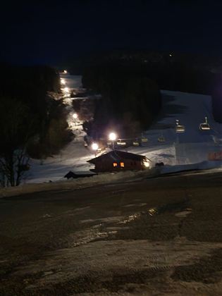Eine verschneite Piste bei Nacht mit beleuchteten Lifts. Im Vordergrund ist eine kleine Hütte zu sehen.