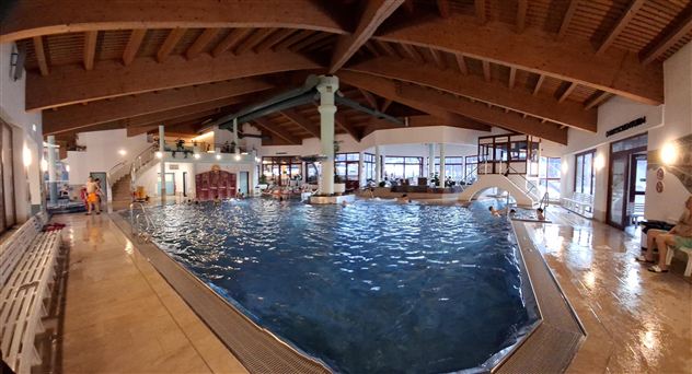 Ein großes Hallenbad mit klarem Wasser und Holzdecke. Umgeben von gemütlichen Liegen und Sitzbereichen für Gäste.