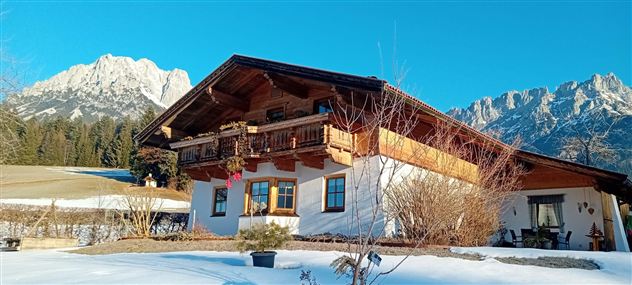 Ein traditionelles Alpenhaus im Schnee mit einem schönen Balkon. Im Hintergrund sind majestätische Berge und ein klarer blauer Himmel zu sehen.