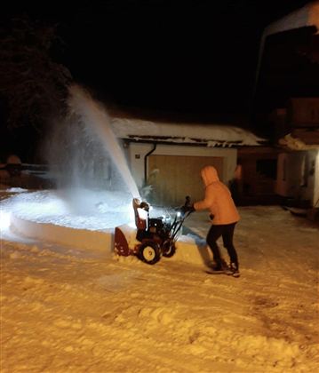 A person is operating a snow blower in a snowy environment at night. The snow is being blown into the air, and a brightly lit house can be seen in the background.