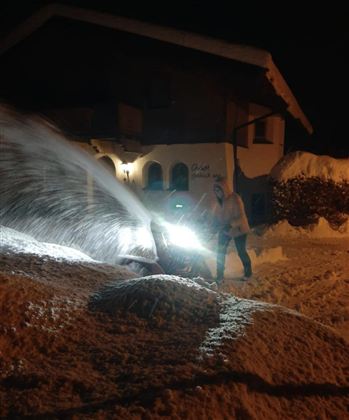 A snowplow is clearing snow at night in front of a house. The snow is being pushed away with bright light.