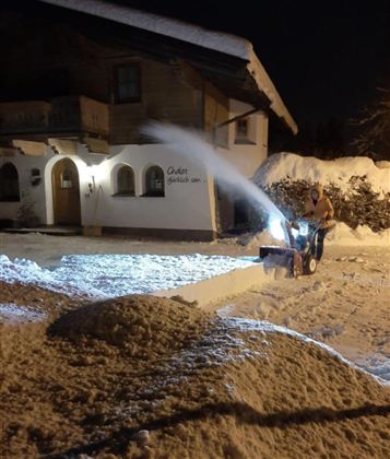 A man is clearing snow with a snow plow at night. In the background, a chalet-like house and a snowy garden can be seen.