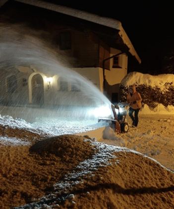 A person is clearing the snow-covered path in front of a house with a snow blower. It is nighttime, and the light from the machine illuminates the surroundings.