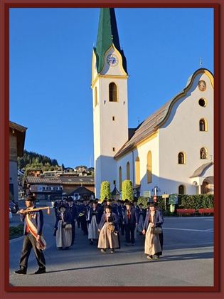 Eine Prozession in traditioneller Tracht geht an einer Kirche vorbei. Die Sonne scheint und der Himmel ist blau.
