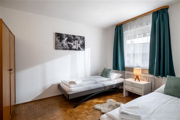 A simple bedroom with two single beds and a bright atmosphere. Large windows and green curtains create a cozy space.