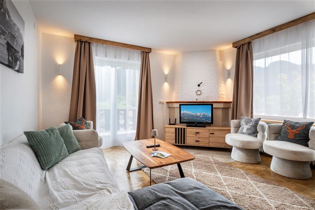 A modern living room with a cozy sofa, a wooden table, and a television. Large windows let in a lot of light and give the room an inviting atmosphere.